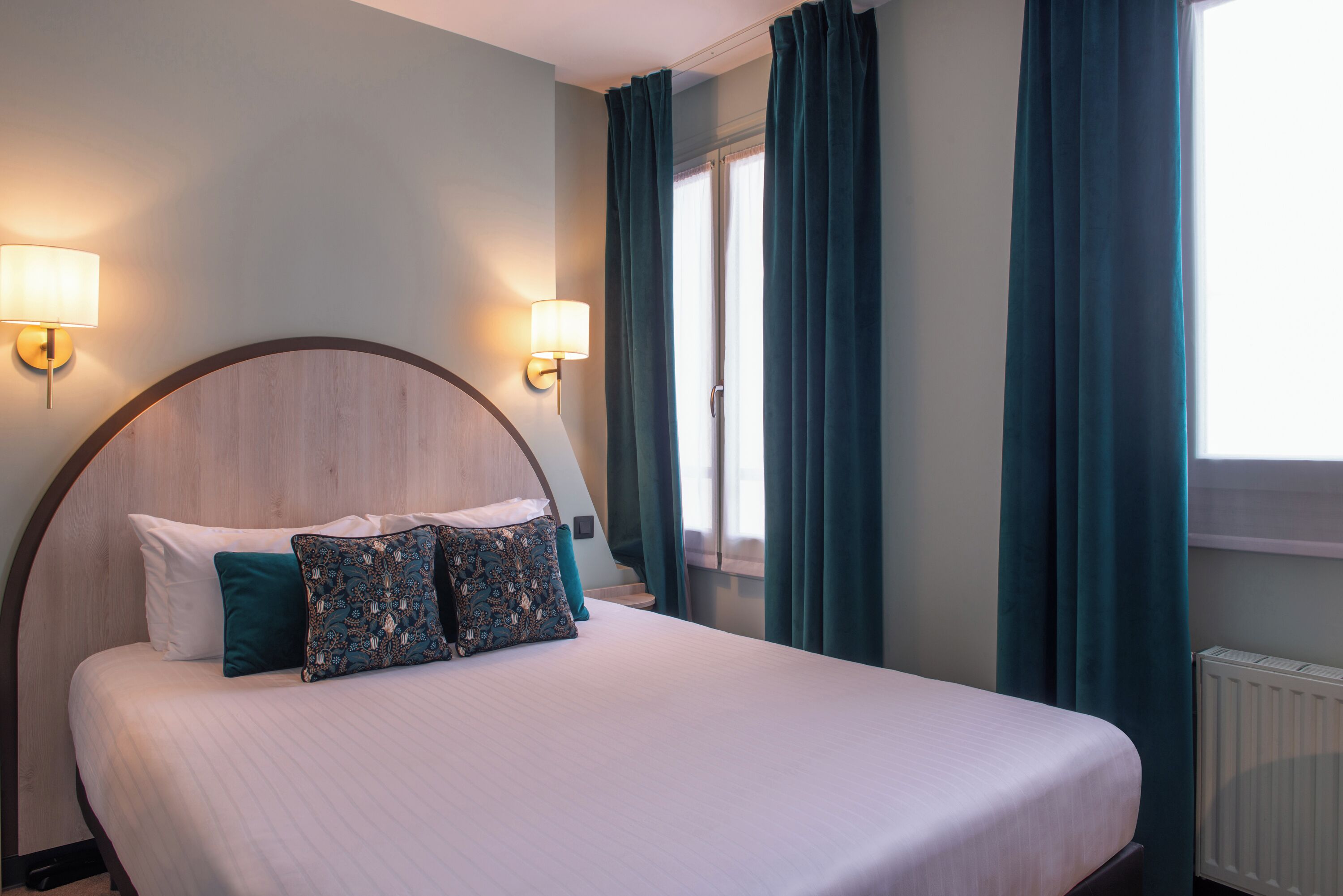 Een opgemaakt bed met een houten hoofdbord, twee wandlampen en teal gordijnen.