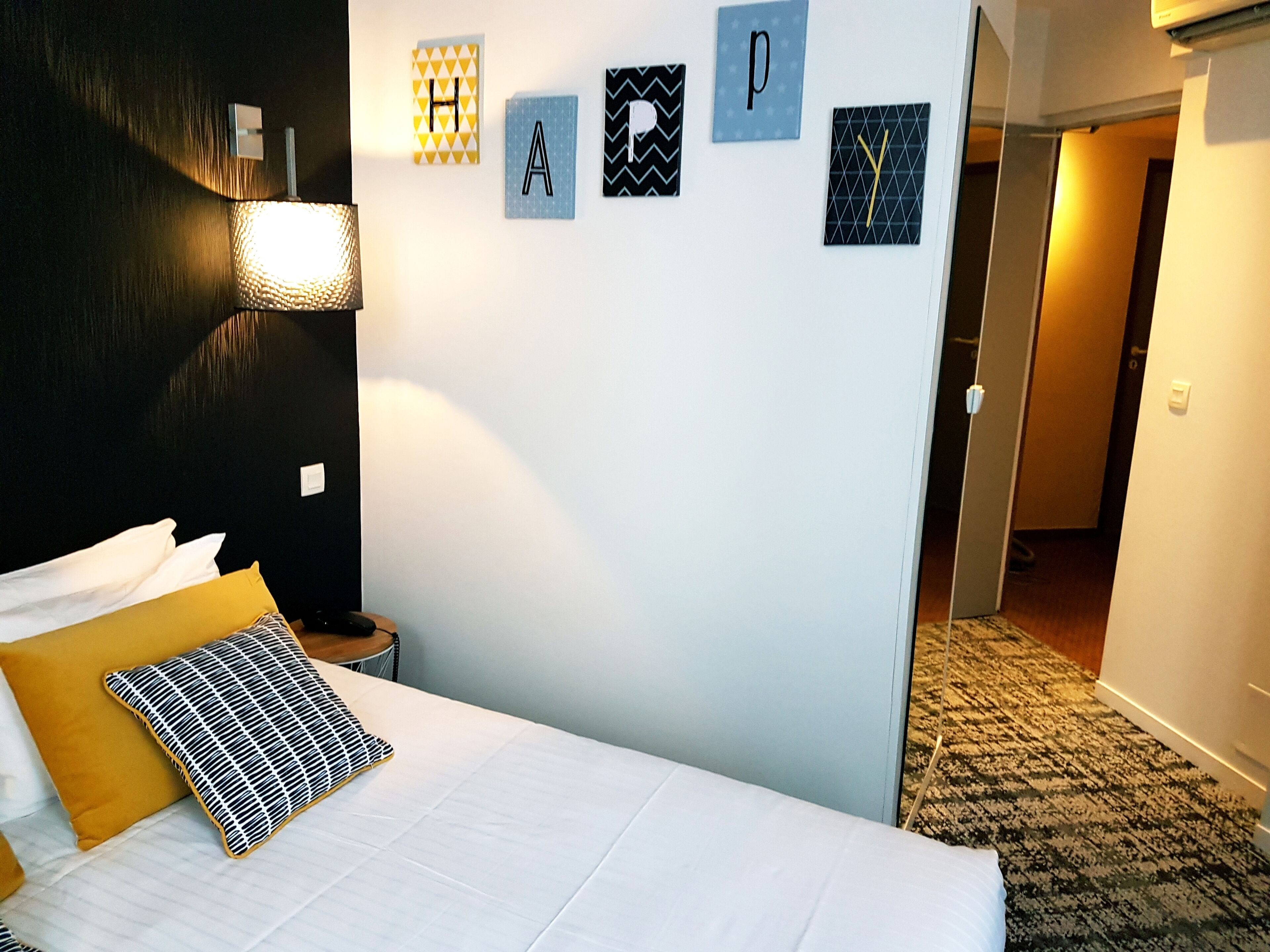 A neatly made bed with a white and mustard-colored duvet, a black and white patterned pillow, and a wall with alphabet letters and geometric patterns.