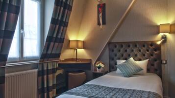 Ein Hotelzimmer mit einem großen Bett, einem Schreibtisch und einem Stuhl. Ein Fenster mit Vorhängen, ein Heizkörper und eine Wandlampe sind vorhanden.