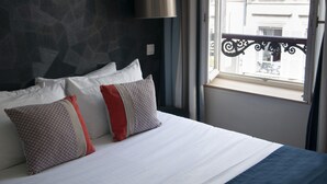 A hotel room with a bed, pillows, a window with a view of buildings, and a balcony railing. - Hotel France Albion (Paris)