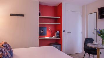 A hotel room with a bed, a desk with a lamp, a coffee machine, and a small cabinet.