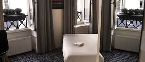 Habitación de hotel con una cama individual, una mesita pequeña y vistas a la calle a través de amplios ventanales.