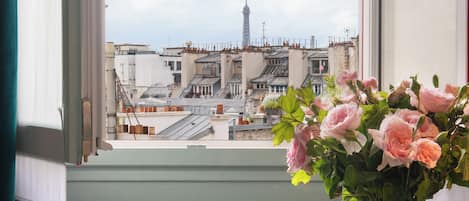 A room with a table, a chair, a vase with flowers, and a view of buildings through a window.
