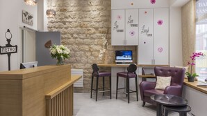 A modern reception area with a stone wall, wooden reception desk, bar stools, a purple armchair, and a computer monitor. - Hotel France Albion (Paris)