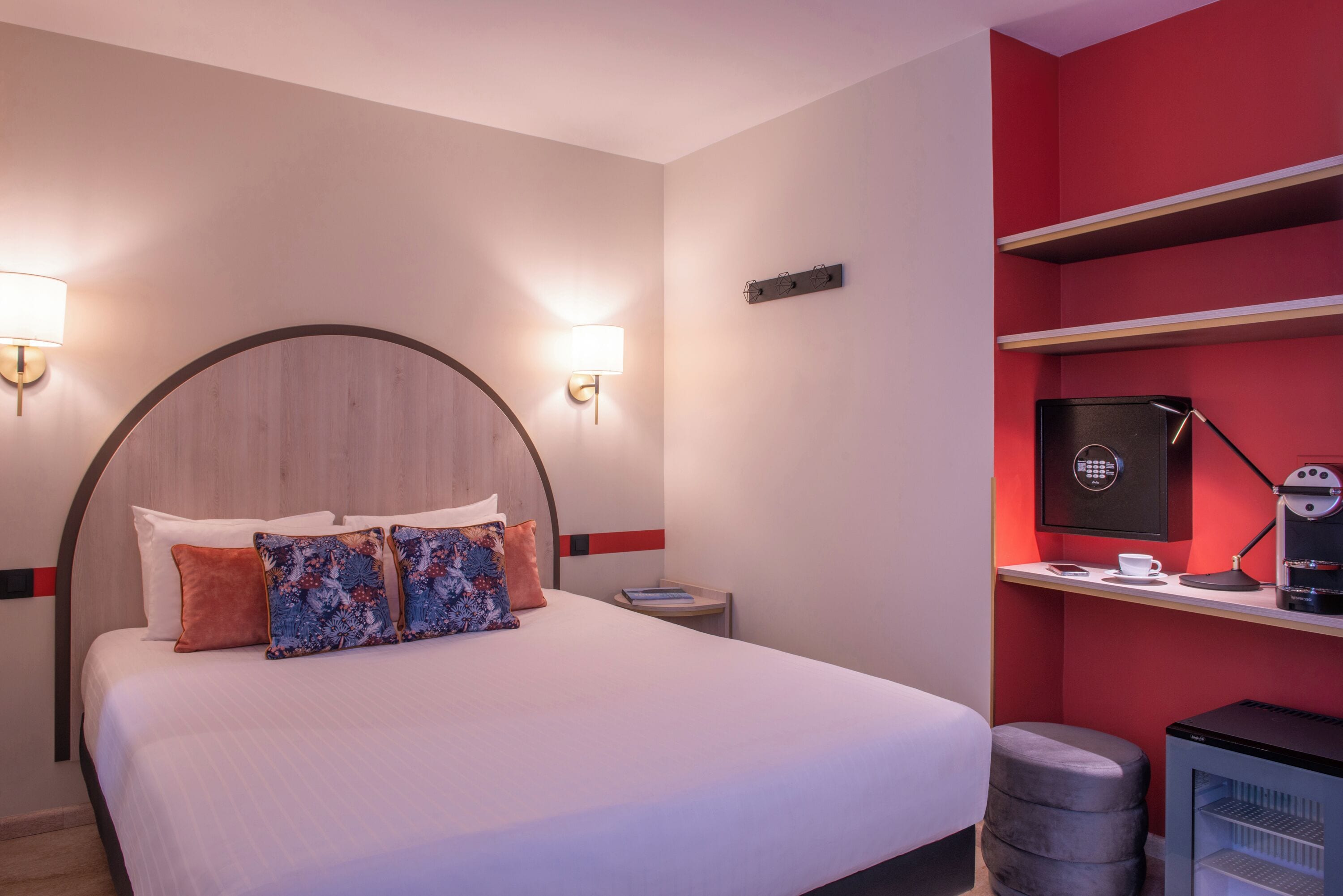Hotelkamer met een bed, twee bedlampen, een nachtkastje, een rode muur en een klein bureau met een koffieapparaat.