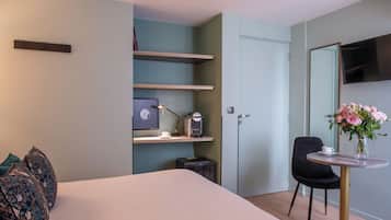 A modern hotel room with a bed, a desk with a coffee machine, a chair, and a vase with flowers.