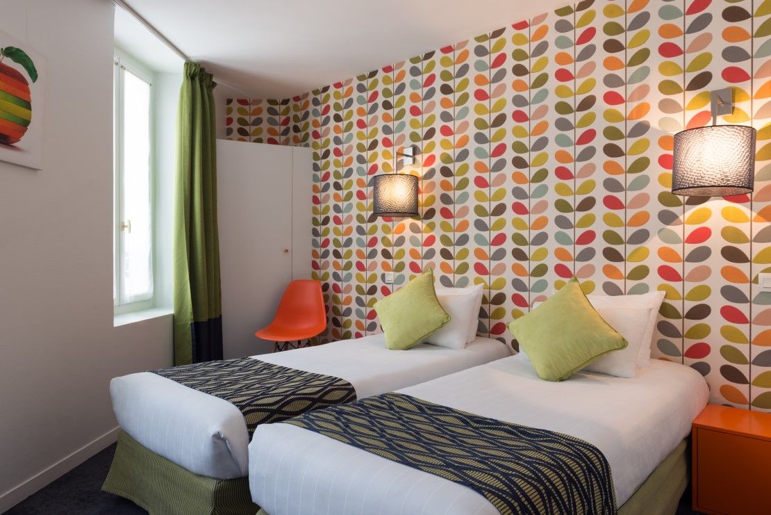 Een hotelkamer met twee bedden, een raam met gordijnen, een stoel en een behang met patroon.