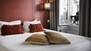 A hotel room with a bed, red and white pillows, a textured throw blanket, a wall-mounted lamp, and a view of the city through a window. - Hotel France Albion (Paris)