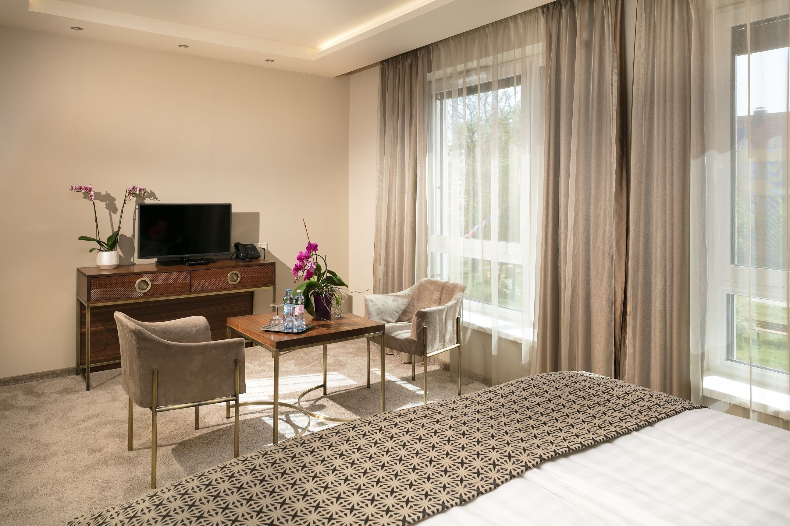 A hotel room with a bed, a desk with a chair, a television, a small table with a vase of flowers, and a large window with curtains.