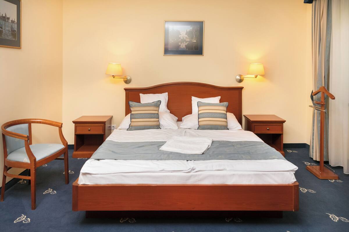 Una cama bien tendida con sábanas blancas y almohadones a rayas, flanqueada por dos mesitas de noche con lámparas, una silla de madera y un cuadro en la pared.
