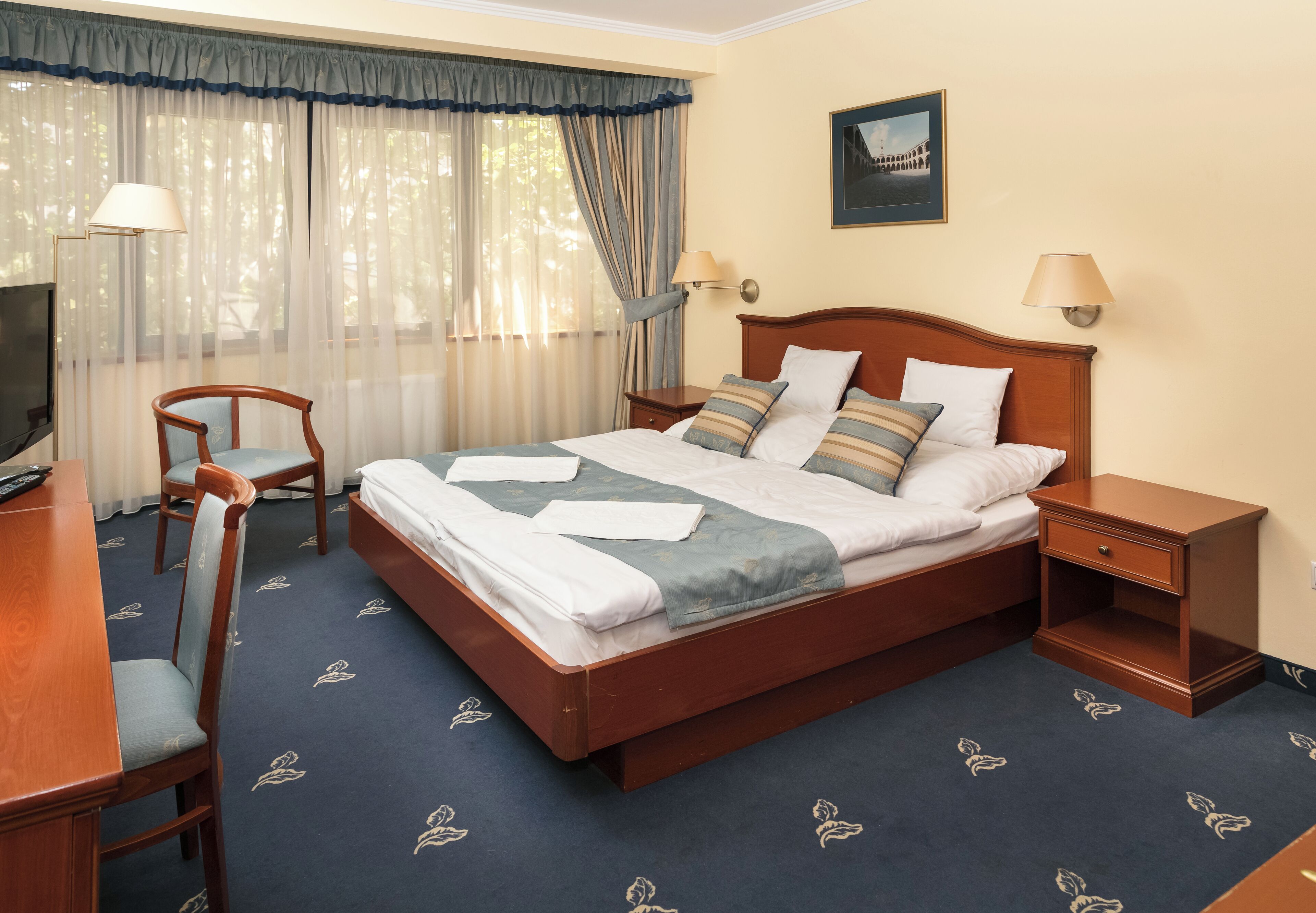 A hotel room with a wooden bed, a desk with a chair, a TV, and two bedside lamps.