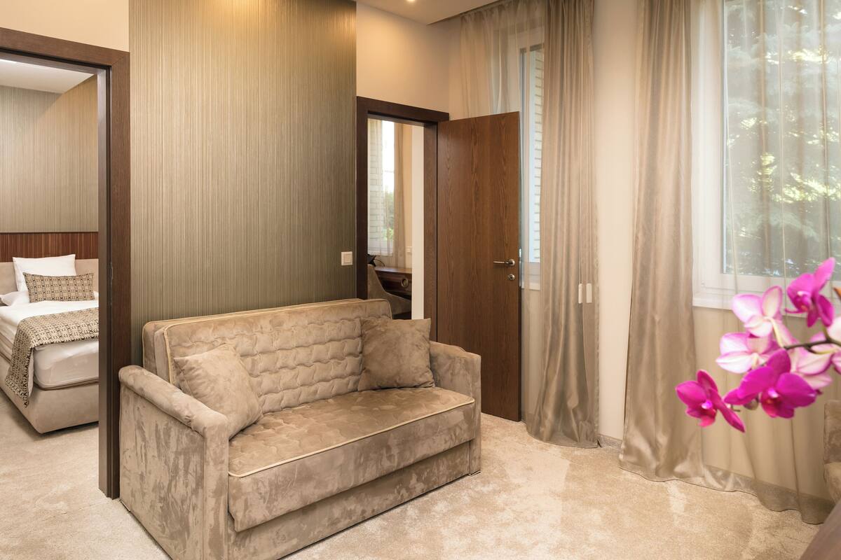 Un dormitorio moderno con un sofá color beige, una puerta de madera y un ventanal con cortinas.