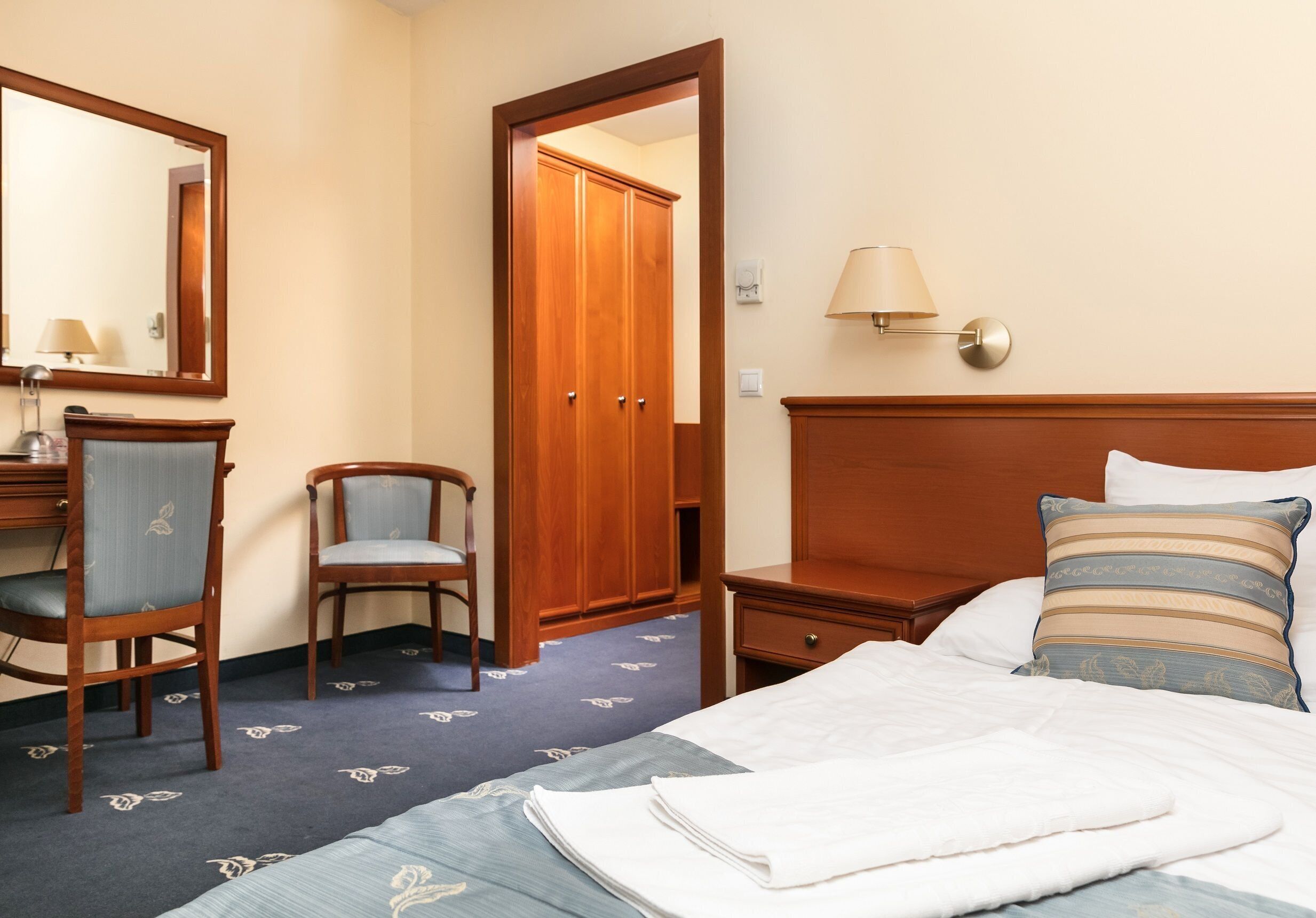 A hotel room with a bed, a nightstand, a lamp, a mirror, a wardrobe, and a chair.