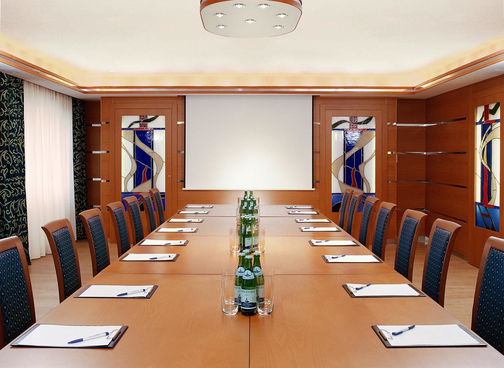 Un salón de conferencias con una larga mesa, sillas, una pantalla de proyector y una botella de agua.