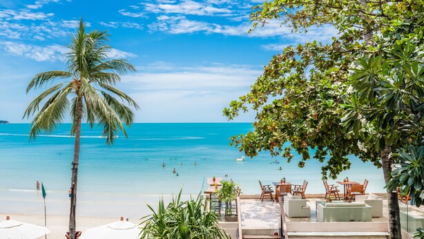 Una playa con agua azul cristalina, palmeras y un área de descanso con mobiliario exterior.
