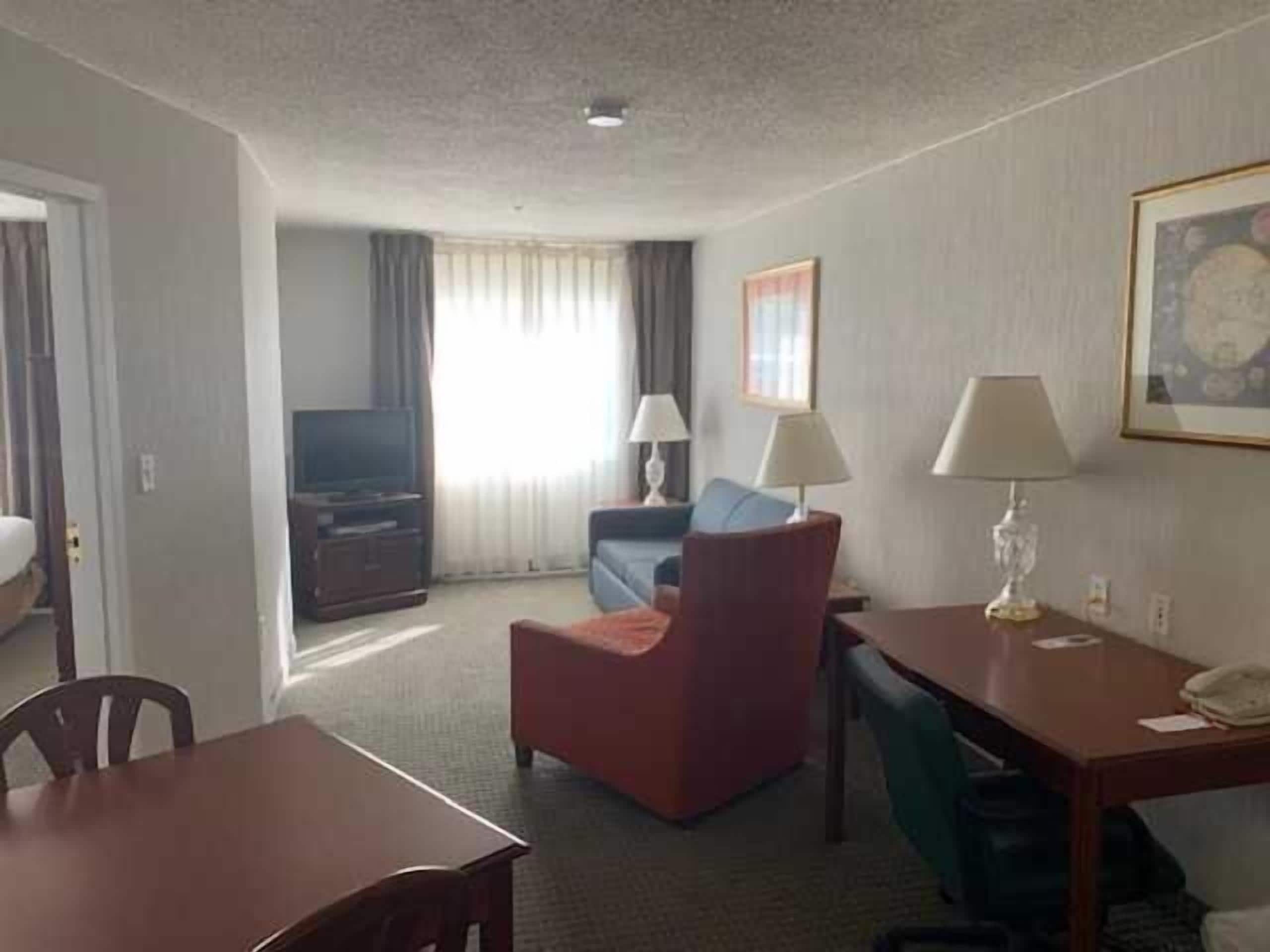Habitación de hotel con cama, escritorio con lámpara, silla, televisor y ventana con cortinas.
