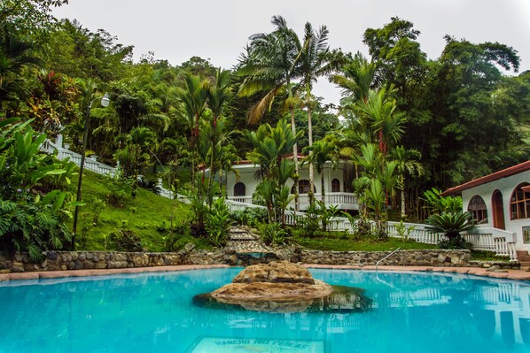 A pool with a rock centerpiece, surrounded by lush greenery and a white building with arched windows. - Rio Perlas Thermal Resort & Spa (Dulce Nombre)