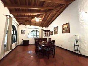 A dining area with a wooden ceiling, white walls, and tiled floor. There is a dining table with chairs, a window with a view, and framed pictures on the walls.