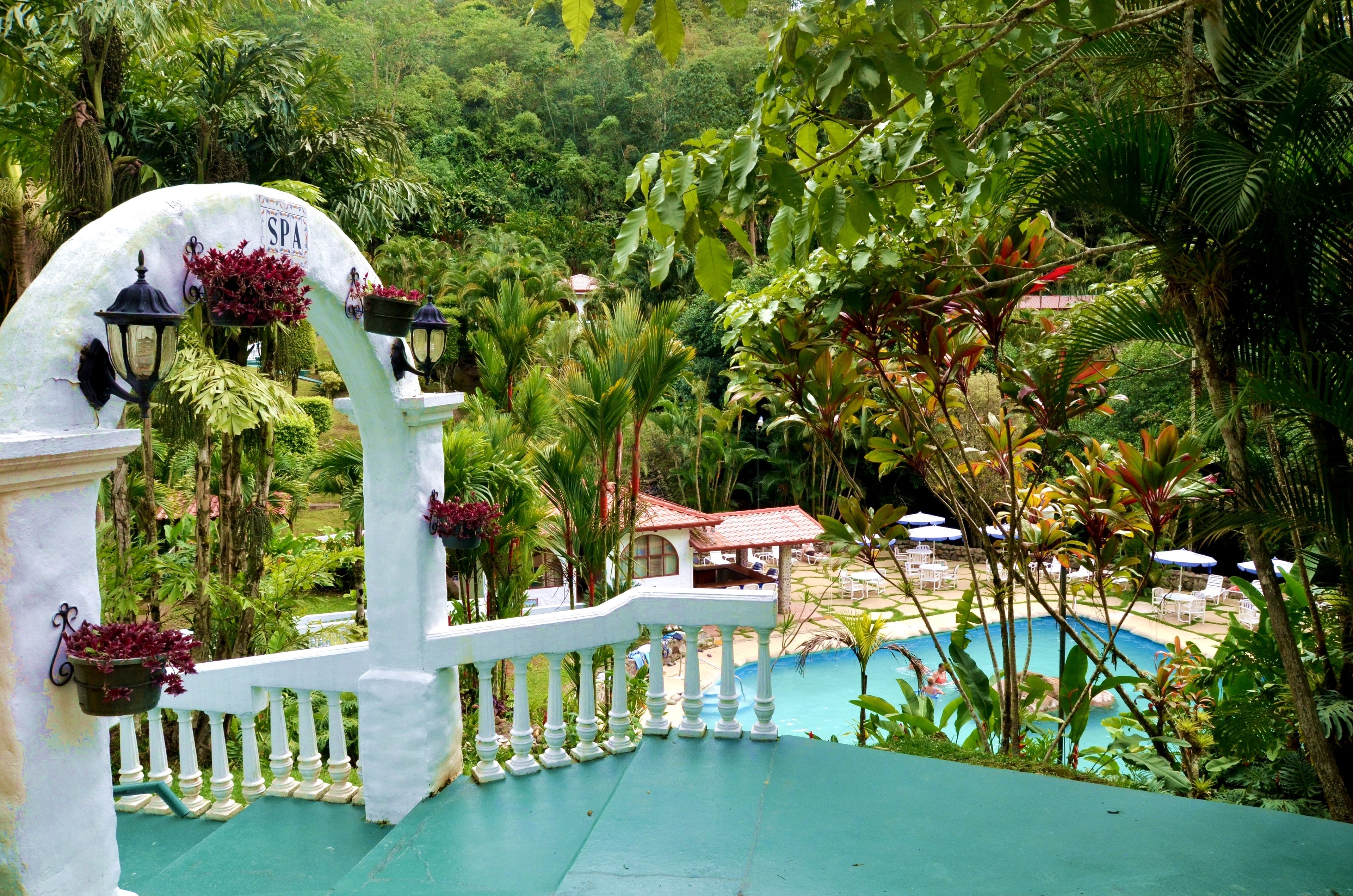 Una entrada arqueada blanca con un letrero que dice “SPA” que da acceso a una zona de piscina rodeada de exuberante vegetación y un pequeño edificio con techo rojo.