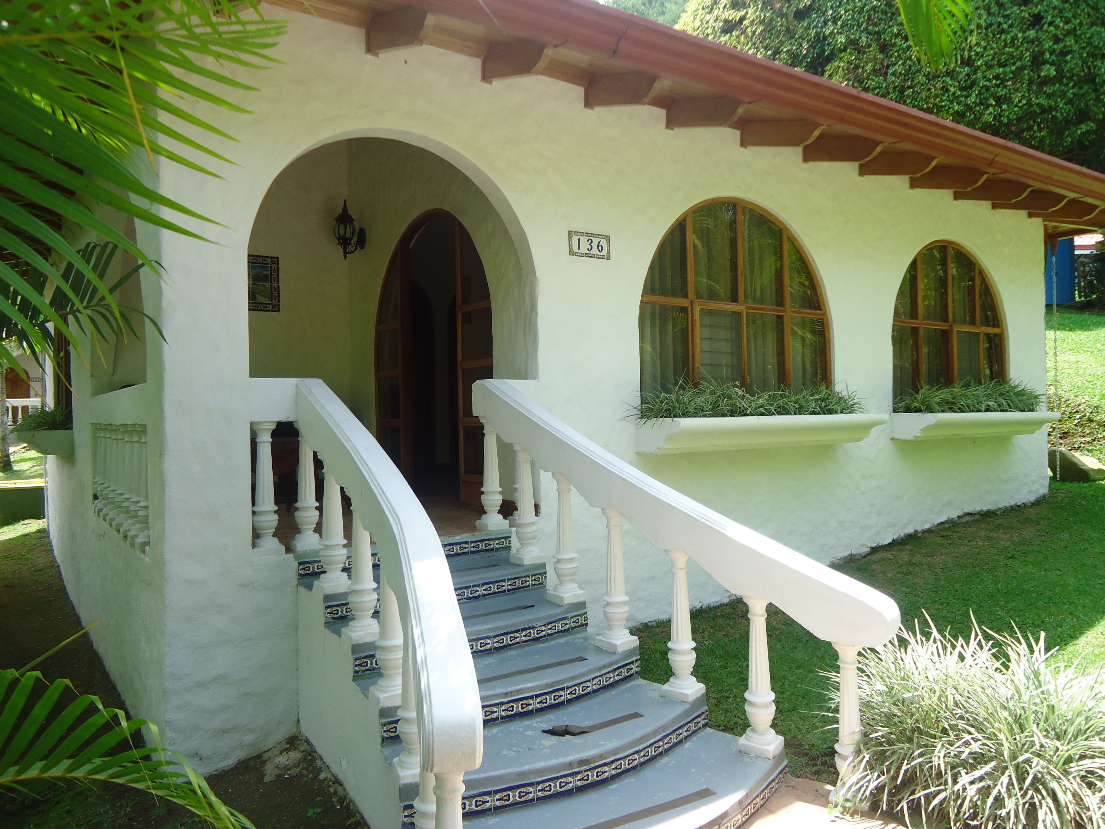 A white building with a staircase leading to an entrance, surrounded by greenery.