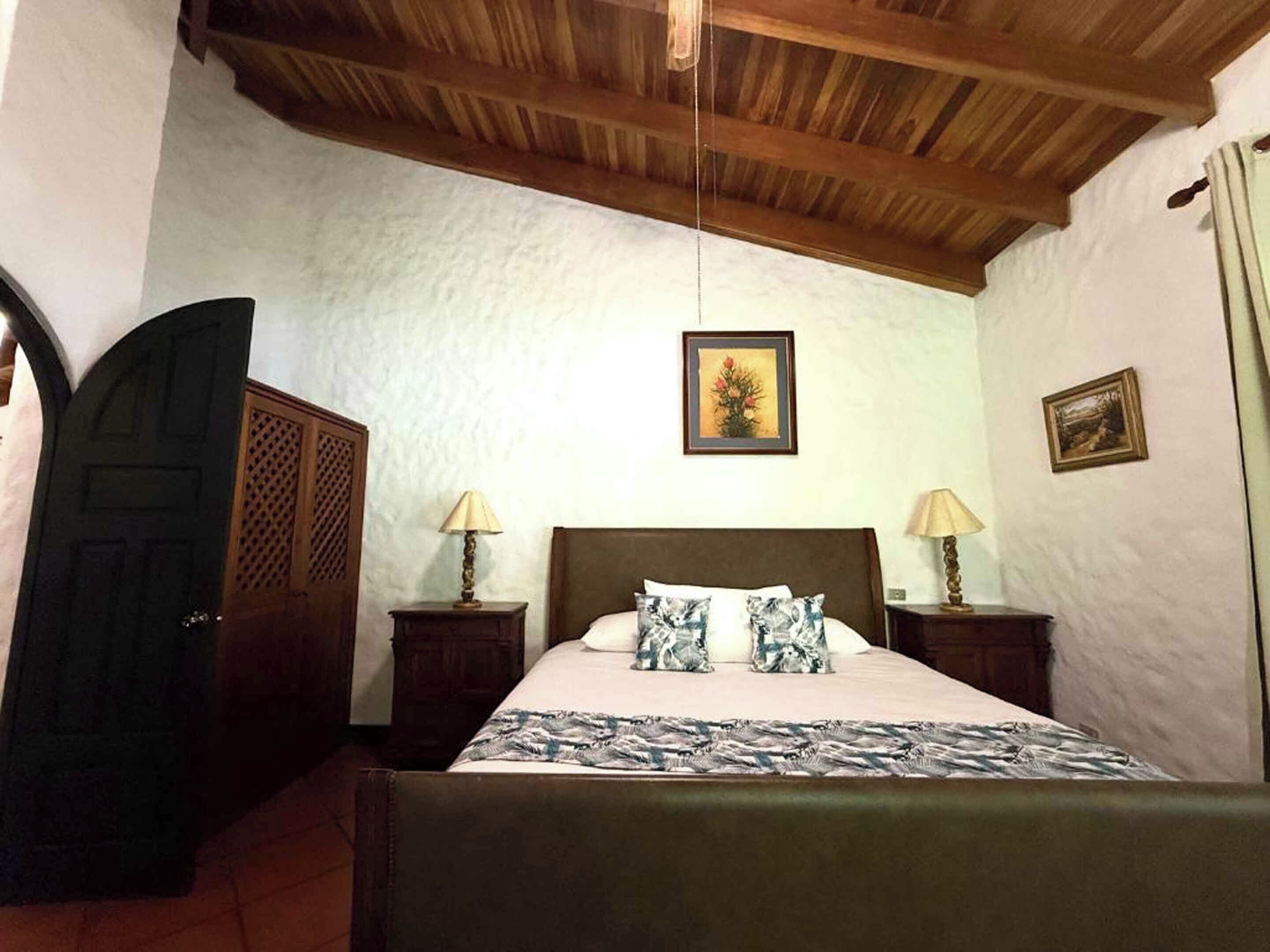 A bedroom with a wooden ceiling, a bed with patterned pillows, two bedside lamps, and a door with a lattice design.