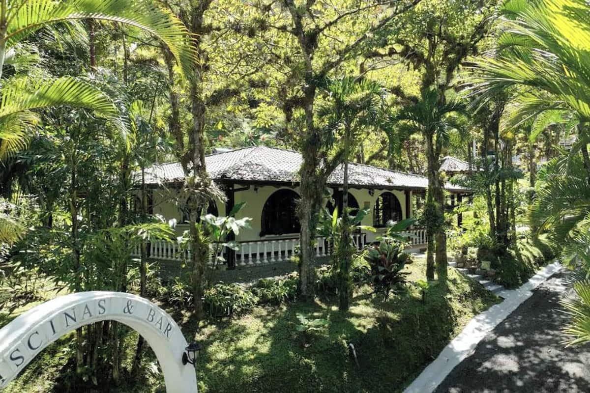A lush, tropical garden with a traditional building featuring arched windows and a thatched roof. A pathway leads to the building, and a sign reads "ESCINAS BAR".