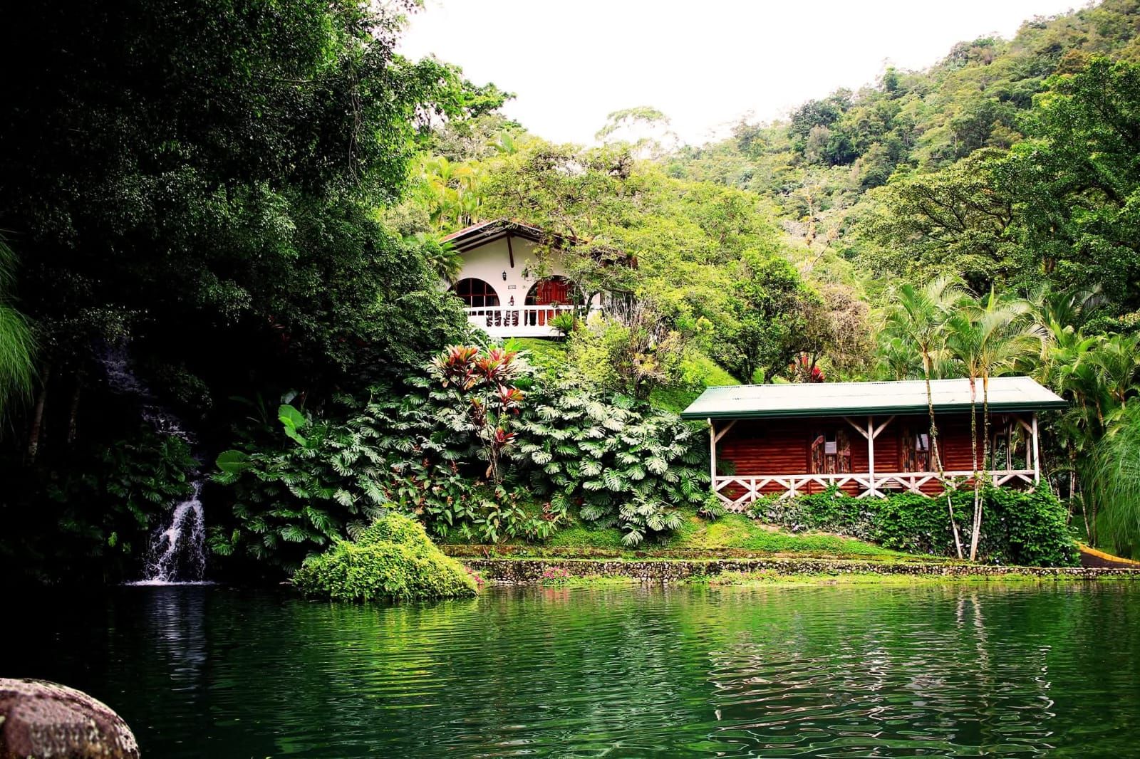A serene lakeside setting with a waterfall, lush greenery, and a small building with a porch.