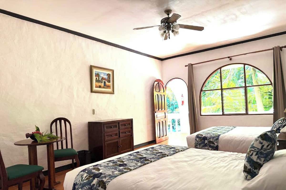 A hotel room with two beds, a desk with a chair, a small table with a vase, a ceiling fan, and a large window with a view of greenery.