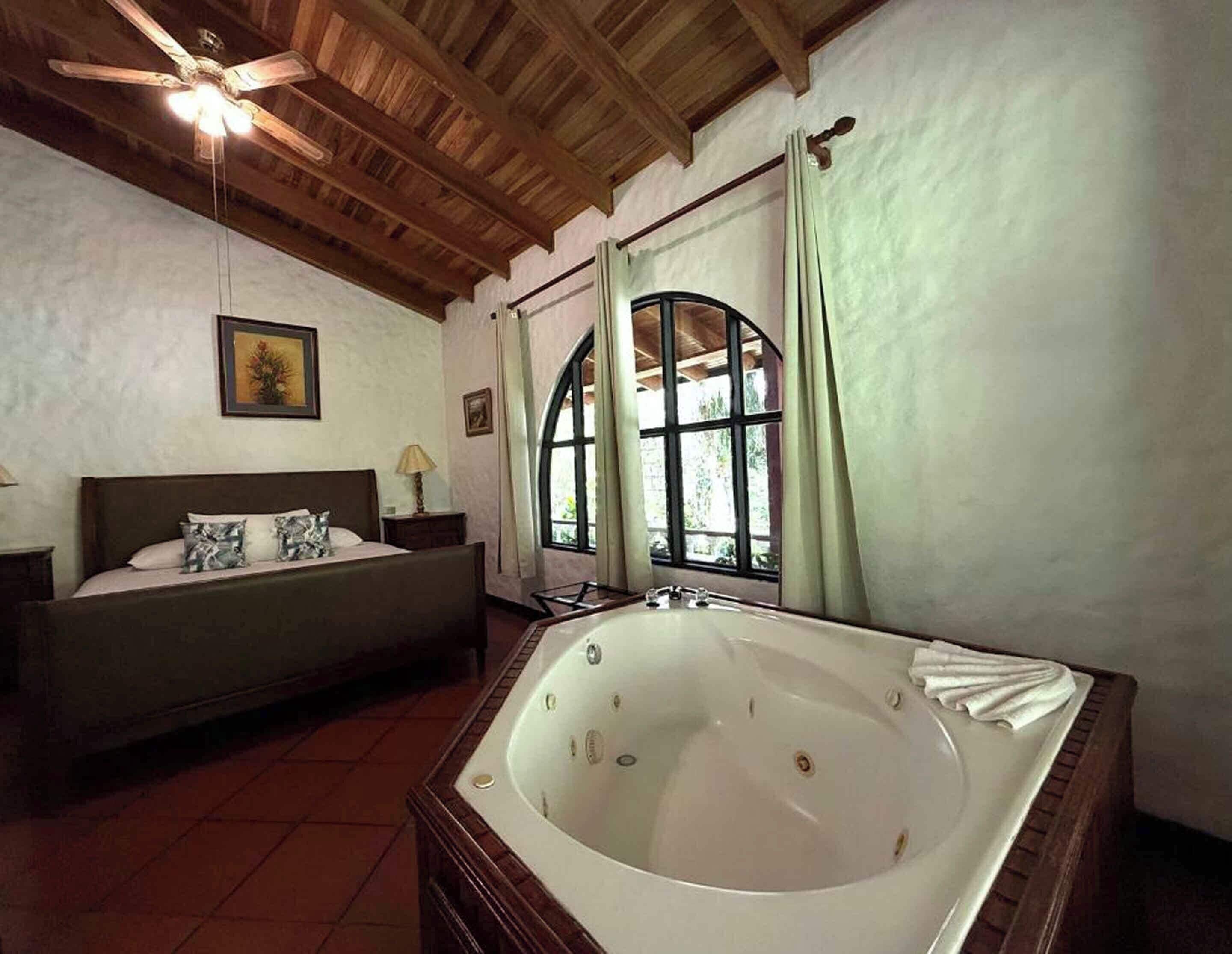 A bedroom with a large bed, a wooden ceiling, a window with curtains, and a bathtub.