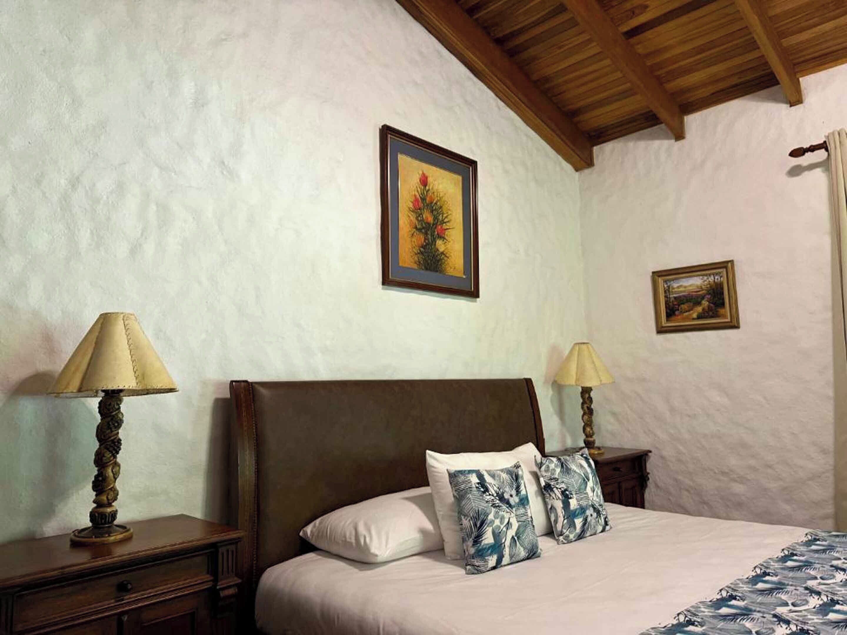 Un dormitorio con cabecera de madera, dos mesitas de noche con lámparas, cama con ropa de cama blanca y almohadas estampadas, y dos cuadros enmarcados en las paredes.
