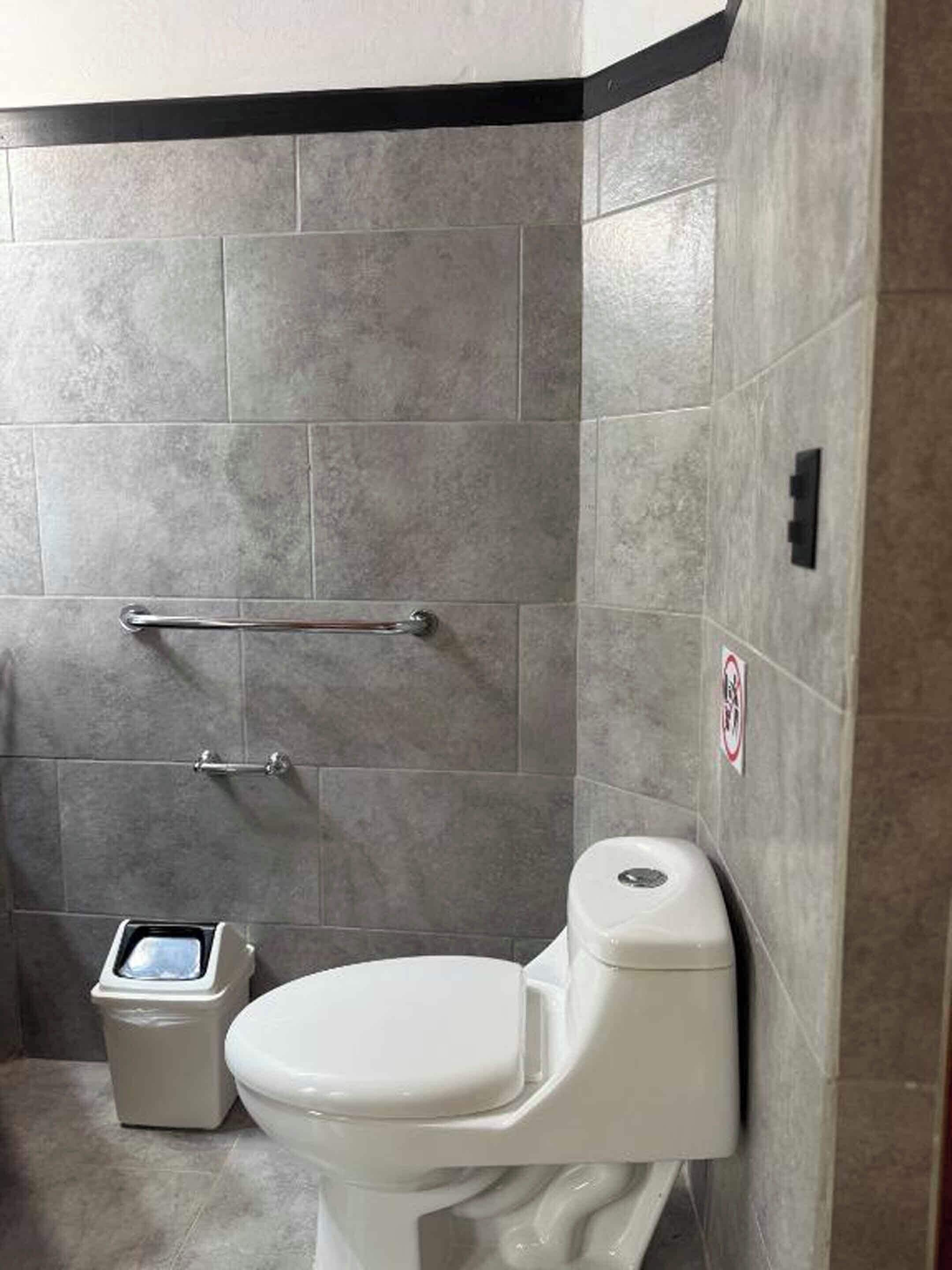 A bathroom with a toilet, a trash bin, and a handrail.
