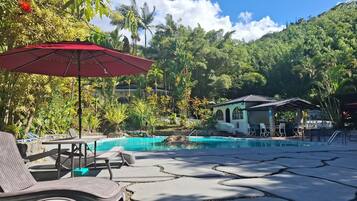 A pool area with lounge chairs and umbrellas, surrounded by lush greenery and a mountain backdrop.