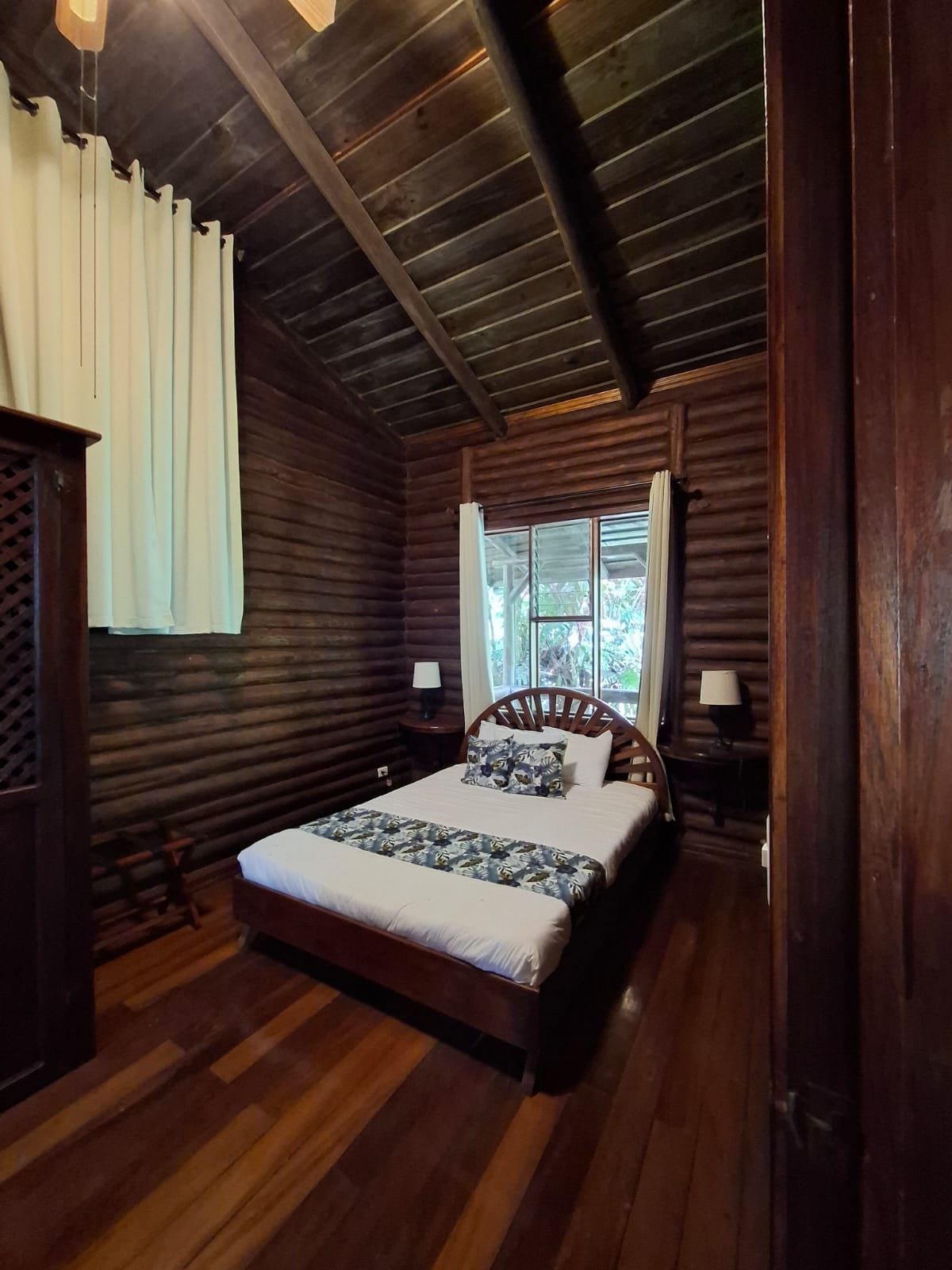 A log cabin bedroom with a wooden bed, two bedside tables, a window with curtains, and a sloped ceiling.