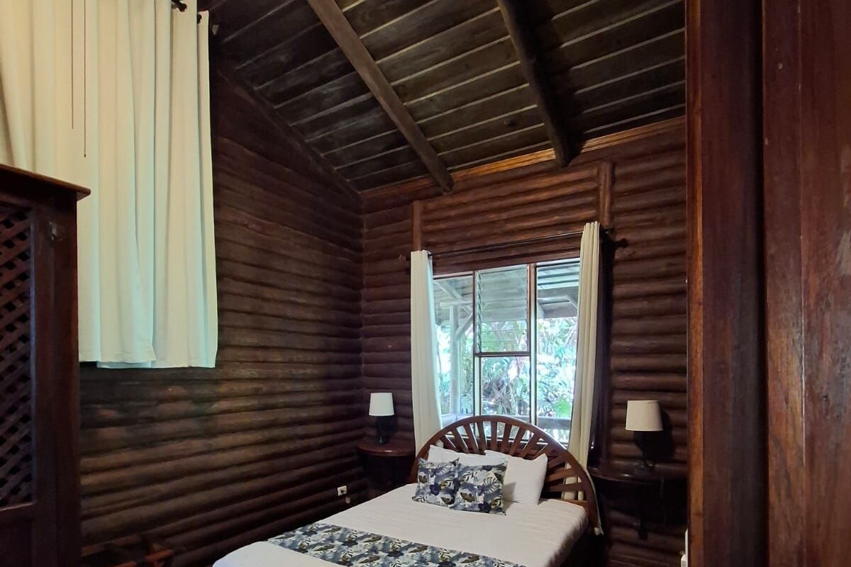 A log cabin bedroom with a wooden bed, two bedside tables, a window with curtains, and a sloped ceiling.