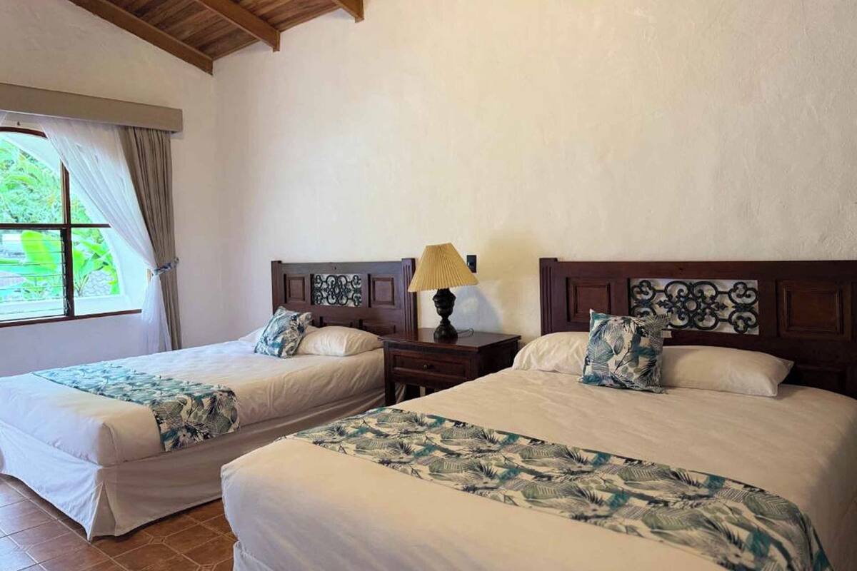A hotel room with two beds, a window with curtains, a wooden ceiling, and a nightstand with a lamp.