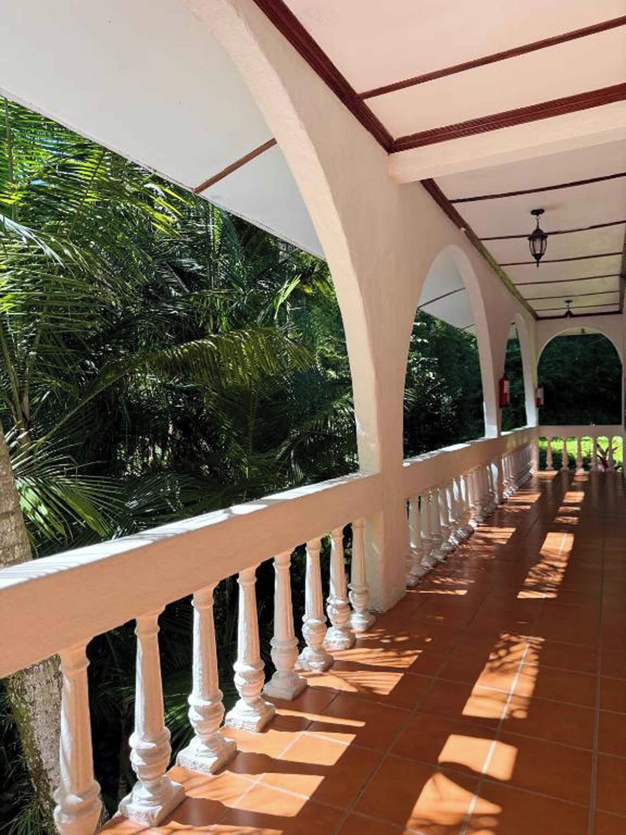A covered corridor with white balustrades and a tiled floor, surrounded by lush greenery.