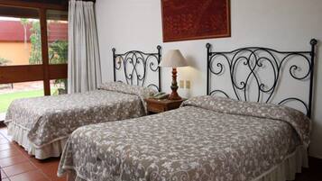 A room with two beds, a nightstand with a lamp, and a window with curtains.