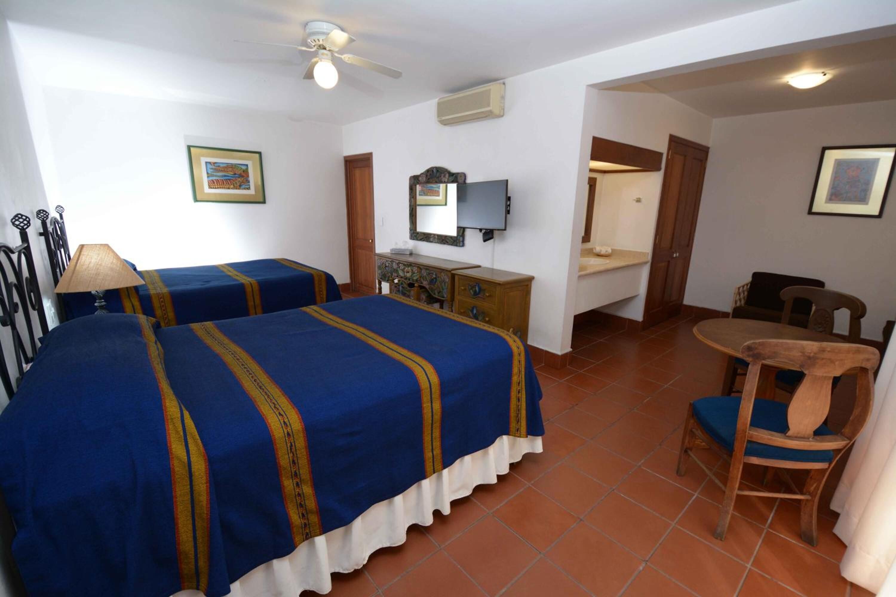 Une chambre d’hôtel avec deux lits, un bureau, une chaise, un ventilateur de plafond et un téléviseur.