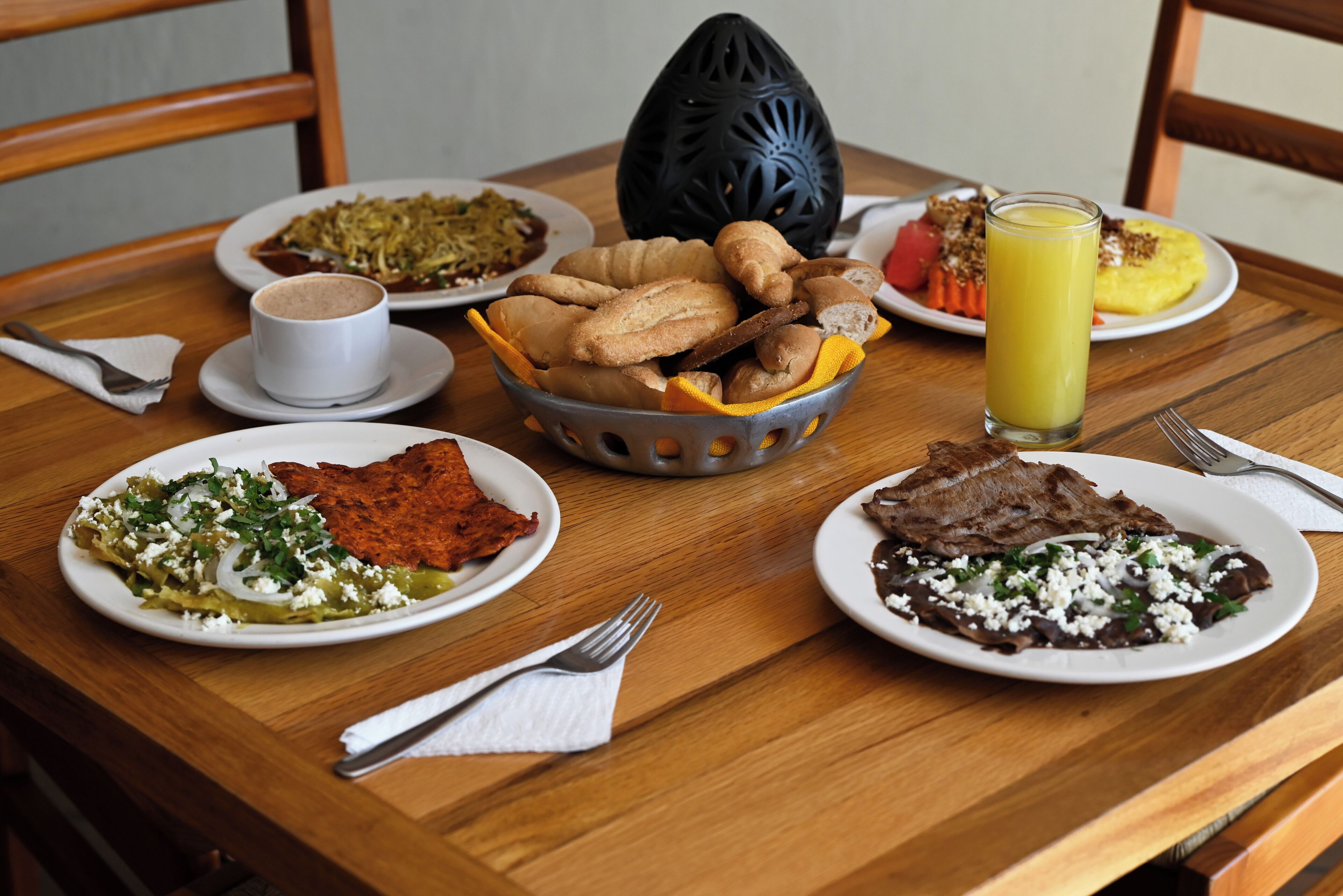 Uma mesa de jantar de madeira com pratos de comida, um copo de suco e um objeto decorativo preto.