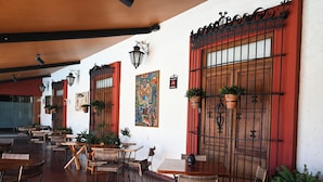 Un patio exterior con mesas y sillas de mimbre, plantas en macetas y una puerta de hierro decorativa.