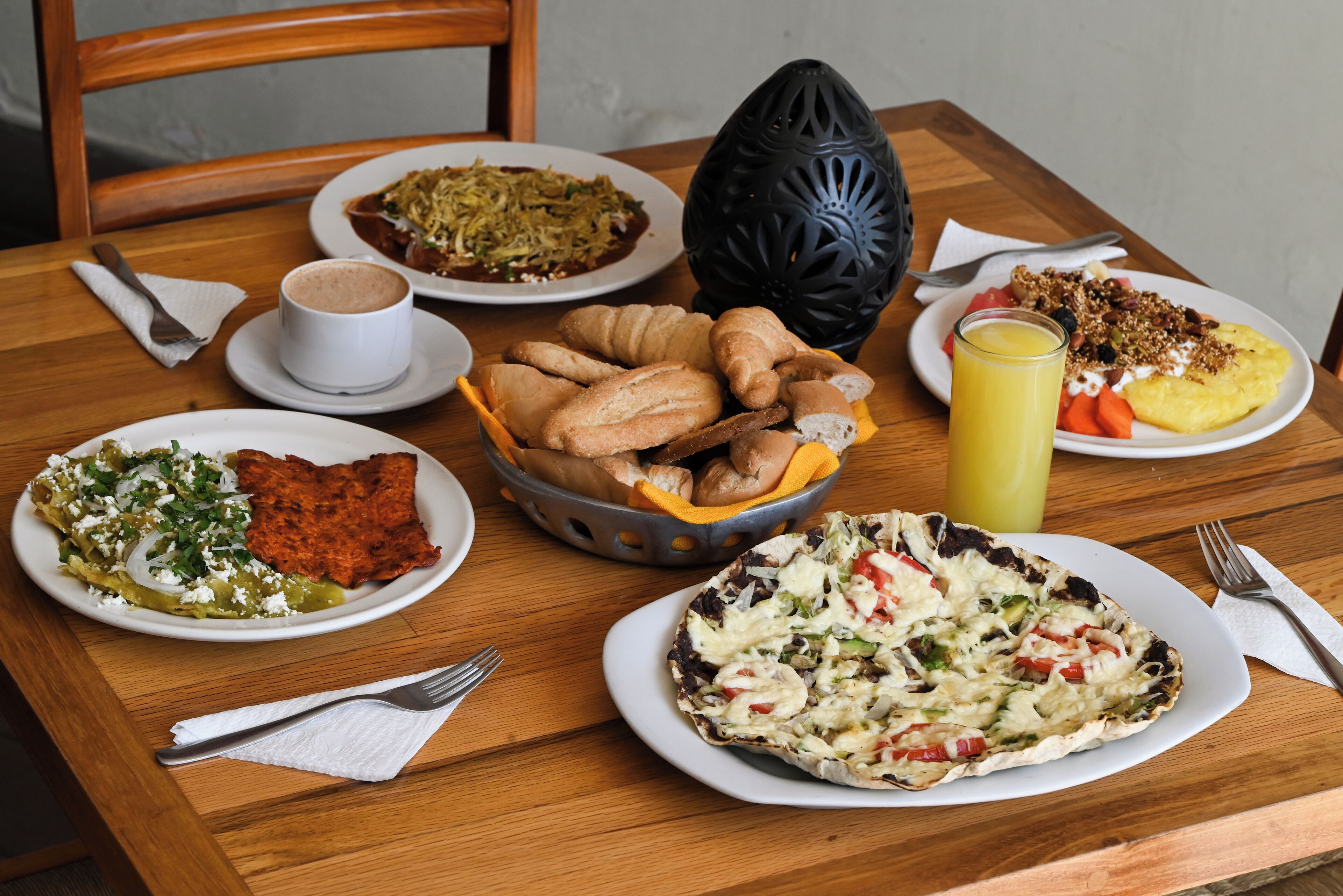 A wooden table with various dishes including a croissant basket, a plate of enchiladas, a plate of eggs, and a glass of orange juice.
