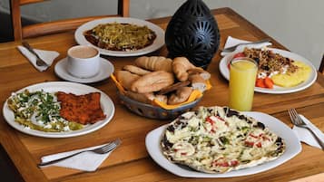 Une table en bois sur laquelle on trouve divers plats, notamment un panier de croissants, une assiette d’enchiladas, une assiette d’œufs et un verre de jus d’orange.