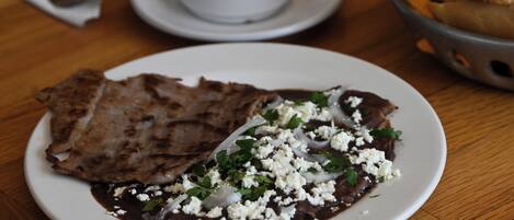 A plate of food with a flatbread, black bean sauce, and crumbled cheese, accompanied by a cup of coffee on a wooden table.