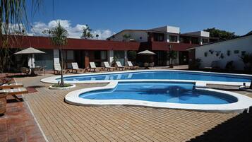 A swimming pool with lounge chairs and umbrellas, surrounded by a brick patio and a building with multiple windows and balconies.