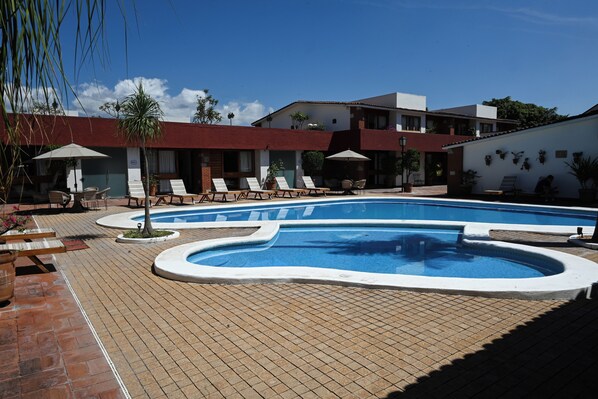 Une piscine entourée d’une terrasse en briques, agrémentée de chaises longues et de parasols, et bordée d’un bâtiment doté de nombreuses fenêtres et de balcons.