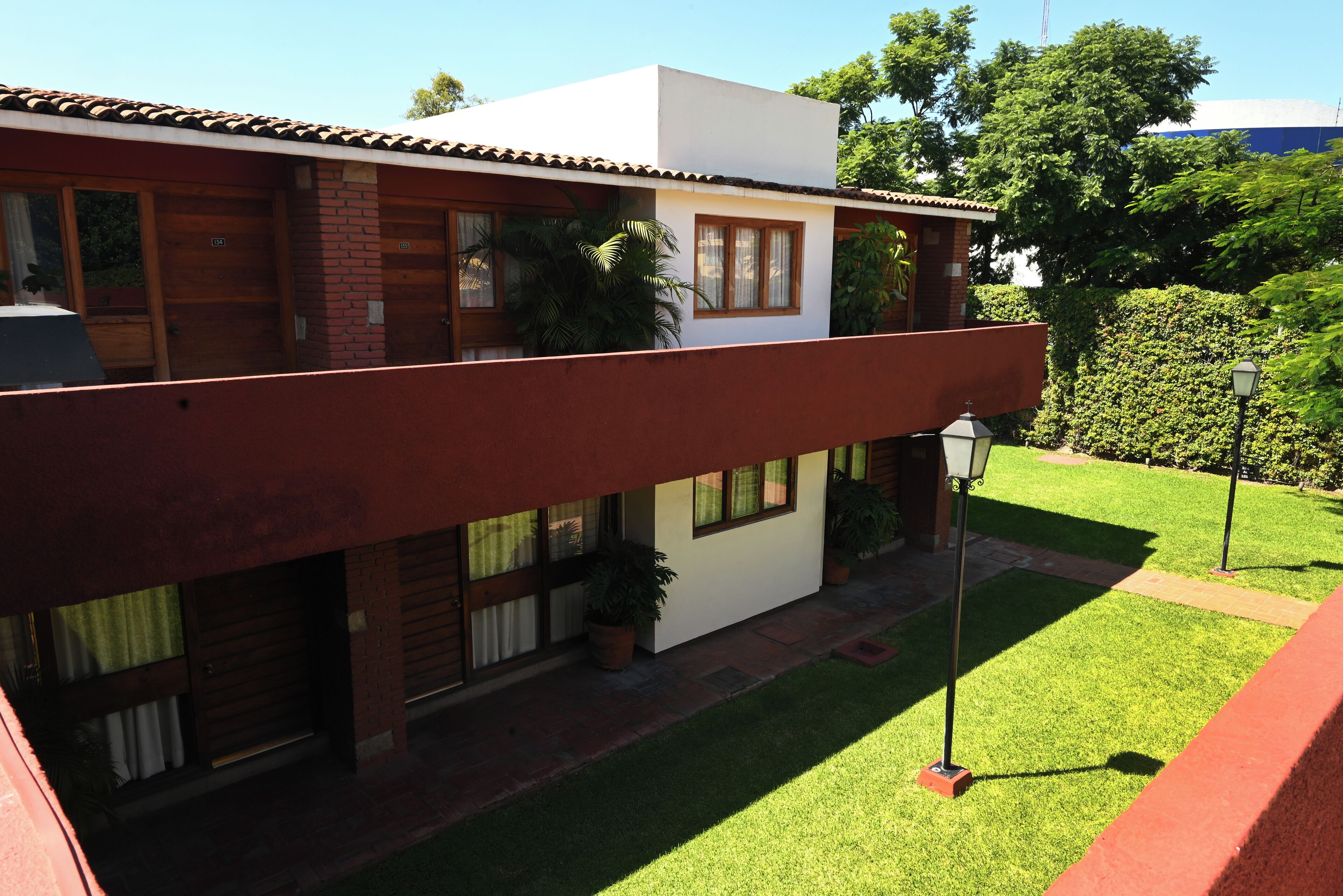 Un edificio de dos pisos con fachada roja, numerosas ventanas y un pequeño jardín con césped y algunas macetas.
