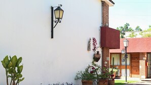 Un edificio con fachada blanca, una chimenea de ladrillo y una puerta de madera. Hay macetas con plantas y un farol en la acera.