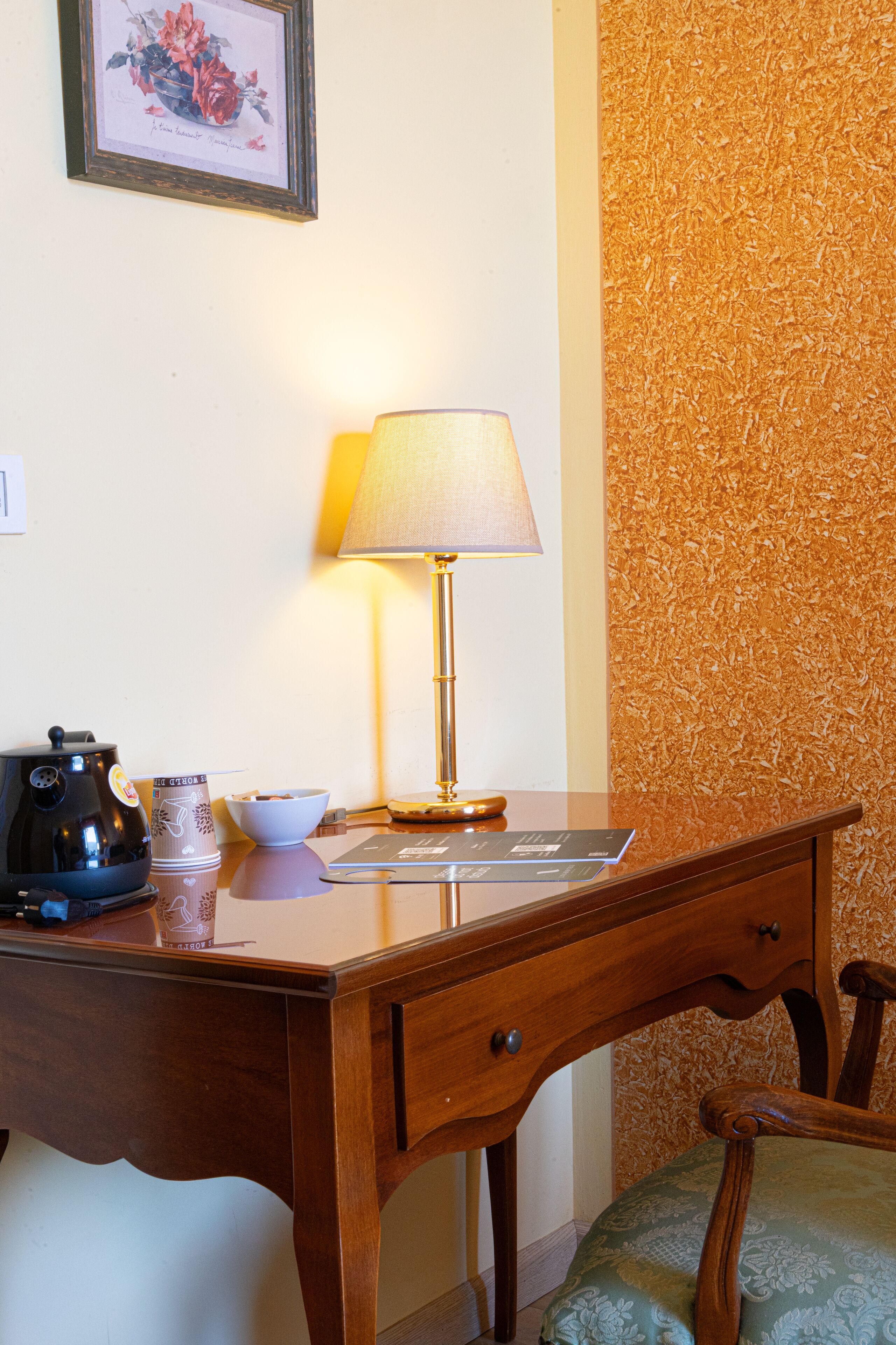 Un escritorio de madera con una lámpara, una cafetera, una taza y un cuadro en la pared.