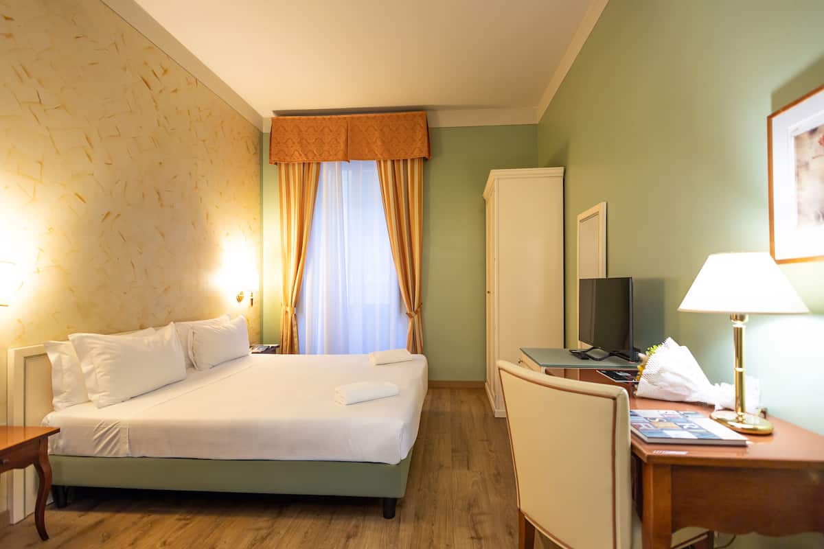 A hotel room with a bed, a desk, a chair, a television, and a lamp.