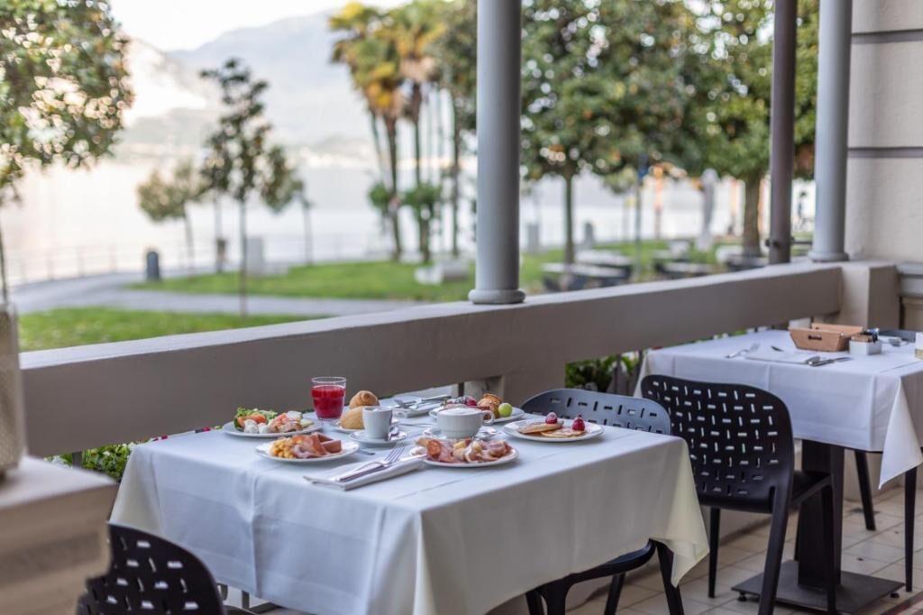 Area pranzo all'aperto con tavoli apparecchiati per la colazione, con vista su uno specchio d'acqua e del verde.